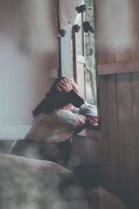 A woman sits indoors gazing thoughtfully out of a window, evoking emotions of loneliness and reflection.